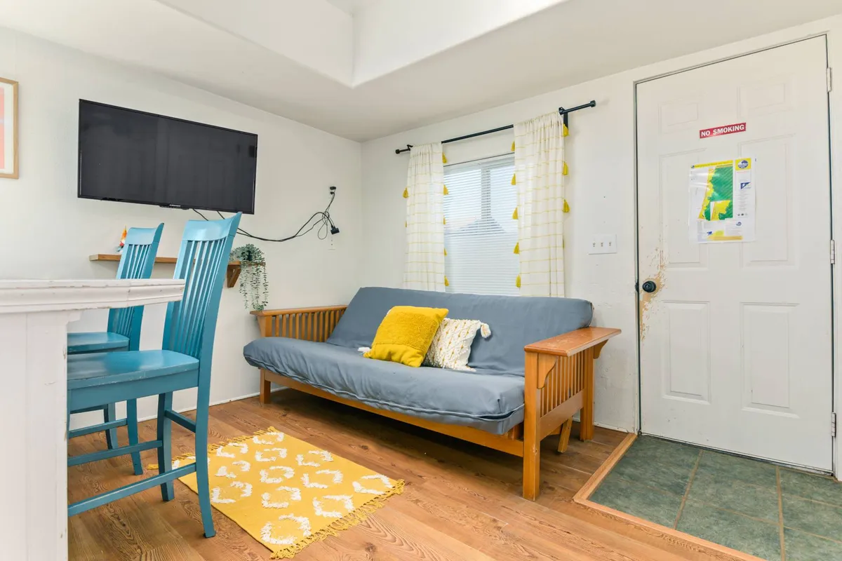 This image centers on a cozy open-plan living area featuring a full futon sofa, vibrant blue chairs, and a cheerful yellow accent rug. A television is positioned for relaxed movie or game nights, and morning sunlight streams through the windows, creating a bright, welcoming atmosphere.