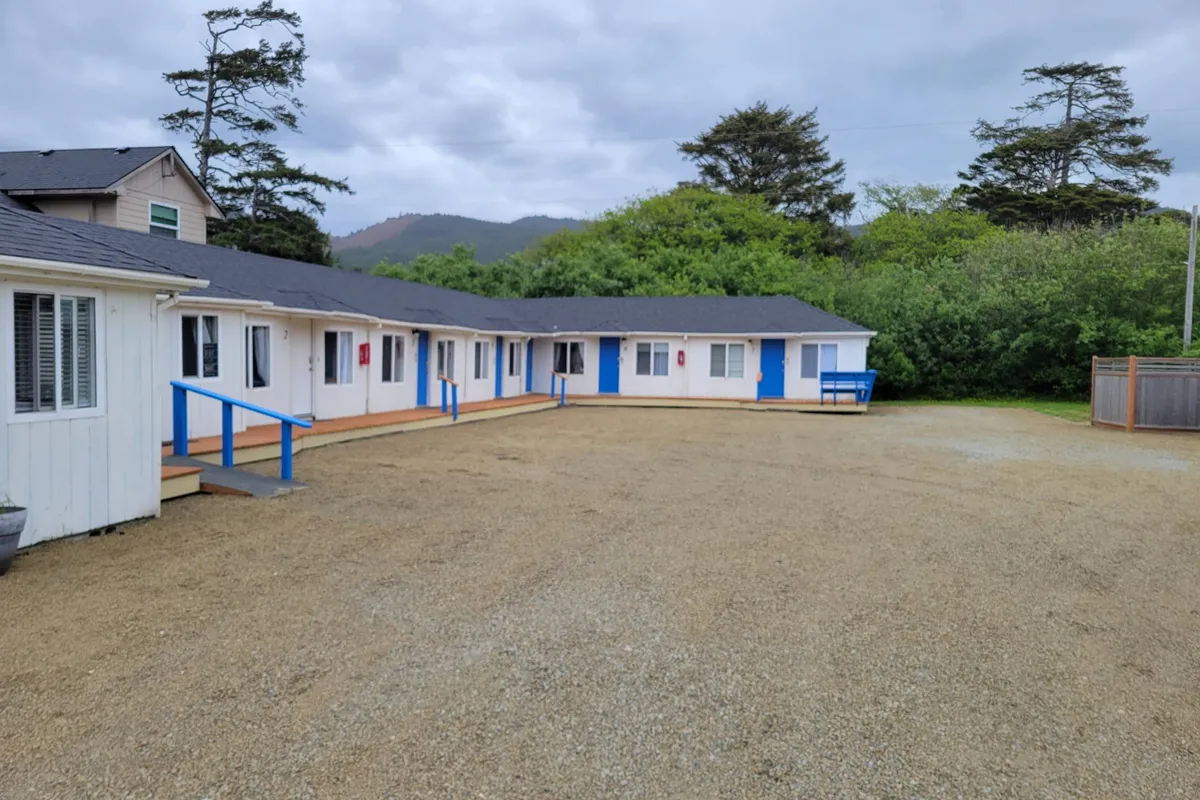 A tranquil gravel courtyard lined with a row of charming cabins featuring blue doors. The space is dotted with outdoor seating and offers plenty of room for guests to relax, socialize, and enjoy the open air.
