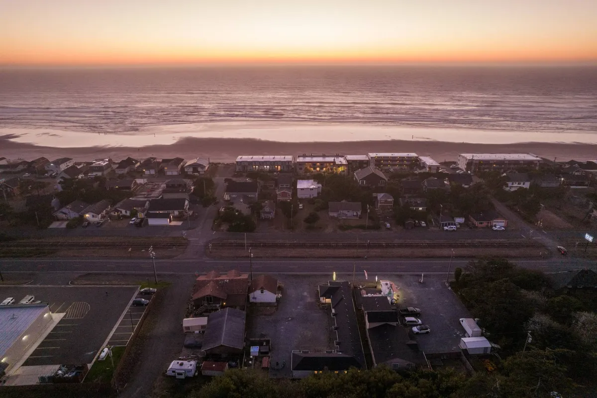A tranquil view shows gentle ocean waves meeting the sandy shoreline, bordered by charming houses and a quiet roadway. The sky glows with colorful sunset hues, signaling the beach’s peaceful atmosphere as day turns to evening.