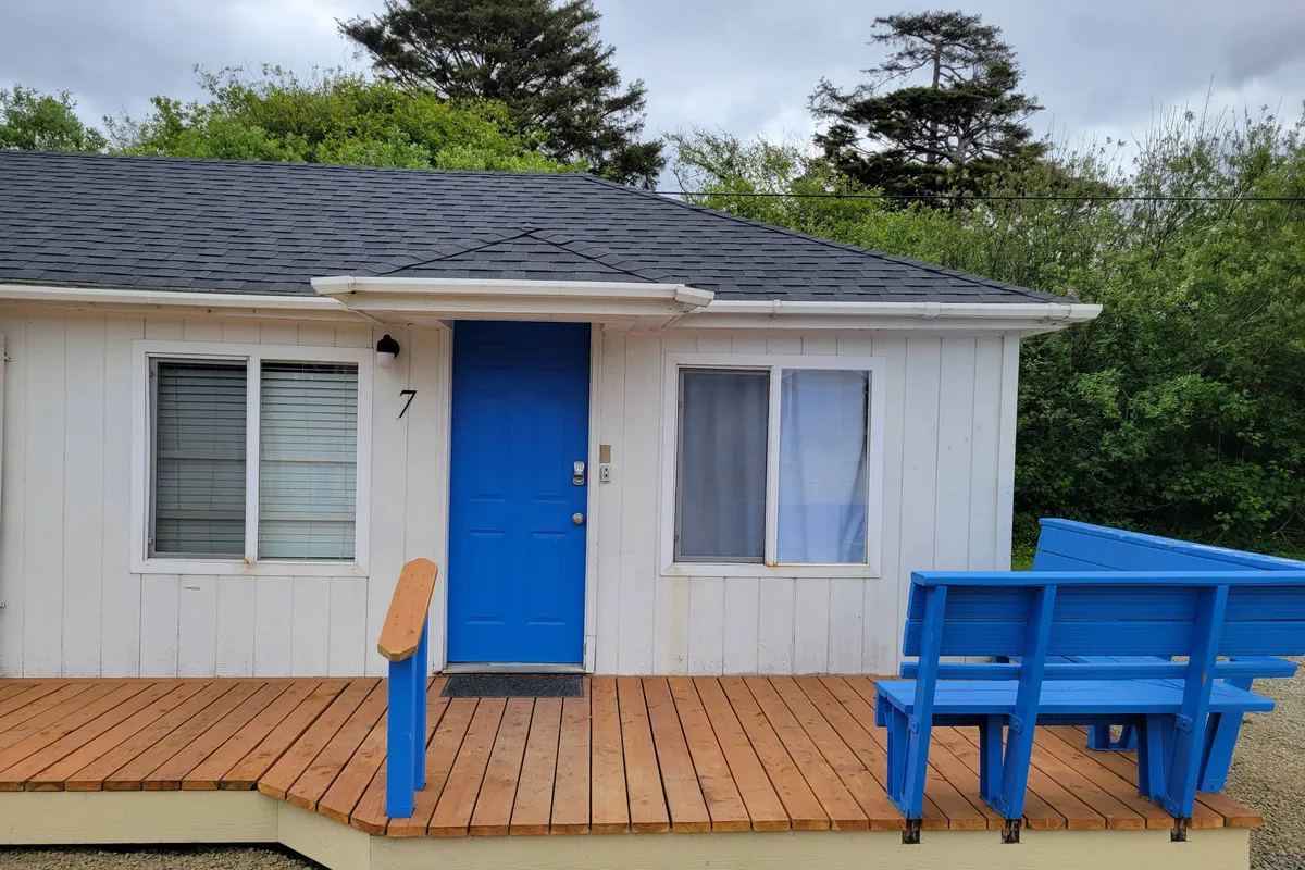 A wooden deck with a blue seating area and a blue door leading indoors. The space is inviting for small gatherings, morning coffee, or casual chats, framed by warm natural light and hints of surrounding nature.