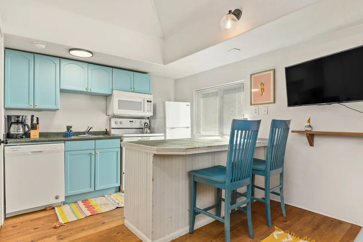 A cheerful kitchen scene with striking blue cabinetry, a central kitchen island, and a warm wooden floor. The kitchen is open to the living area and features space for casual dining and gathering, ideal for cooking together or sharing a casual meal. A TV on the wall connects the kitchen to the living area, reinforcing the open-plan concept.