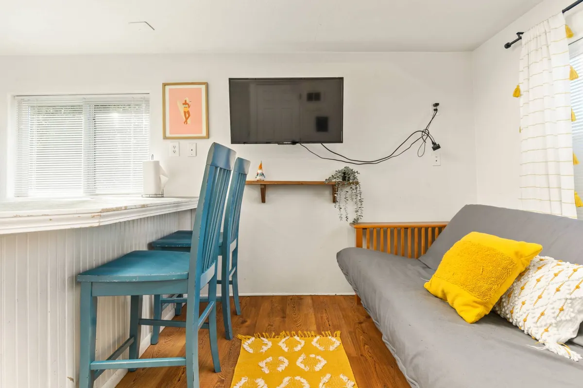 An inviting space that seamlessly blends a sofa with decorative pillows, vibrant yellow accent rug, blue bar stools, and a wall-mounted TV. The area is designed for casual dining, relaxing, and socializing, making it a versatile part of the open-plan hub.
