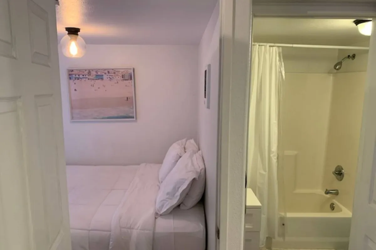 This image features a cozy bed with crisp white linens, a framed artwork of a beach scene above the bed, and the bathroom area visible in the background. Soft natural light fills the room, creating a calm and inviting ambience, while the décor and layout emphasize the room’s relaxing qualities.