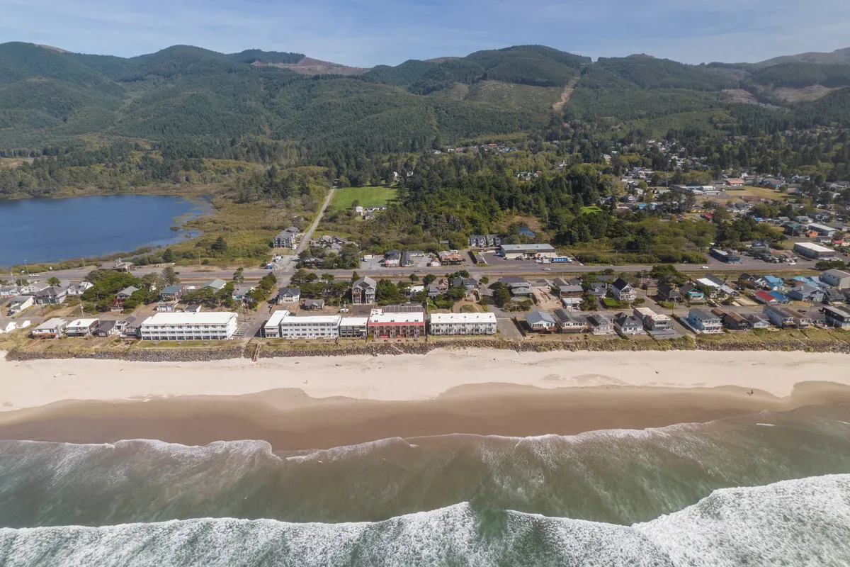 The hero image showcases a sweeping view of Rockaway Beach, with gentle ocean waves rolling onto the sandy shore, the quaint town buildings nestled along the coastline, and a dramatic mountain backdrop rising in the distance. The scene evokes the tranquil charm of a coastal retreat, with opportunities for beachcombing, surfing, and exploring local shops clearly suggested by the landscape.